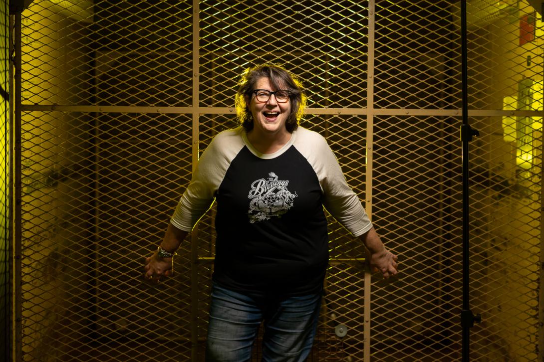A woman is standing in front of a service elevator with her arms reaching behind her, gripping the meshed door. She is dark haired, wearing glasses, a black and white baseball style T-shirt and jeans, and laughing.